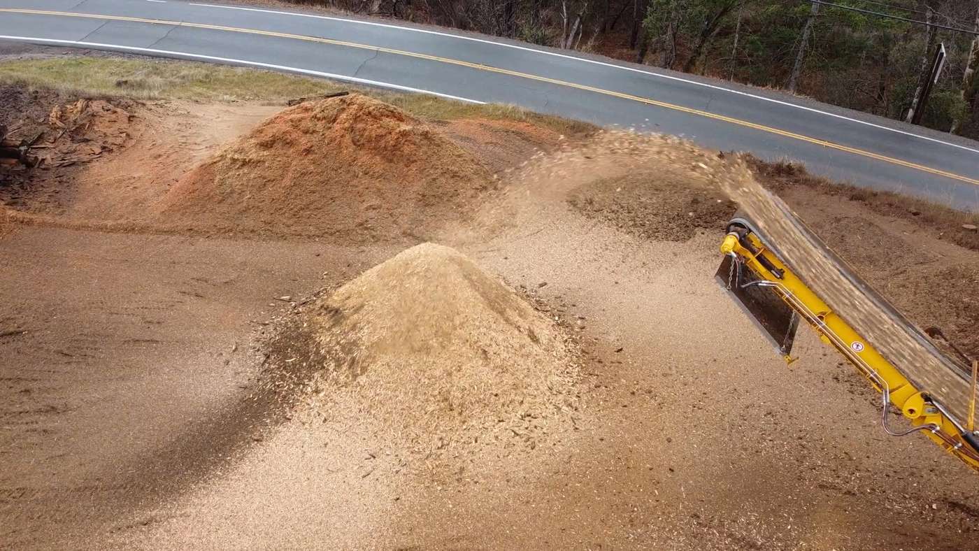 Fresh wood chips pouring off the conveyor onto a growing pile beside the road