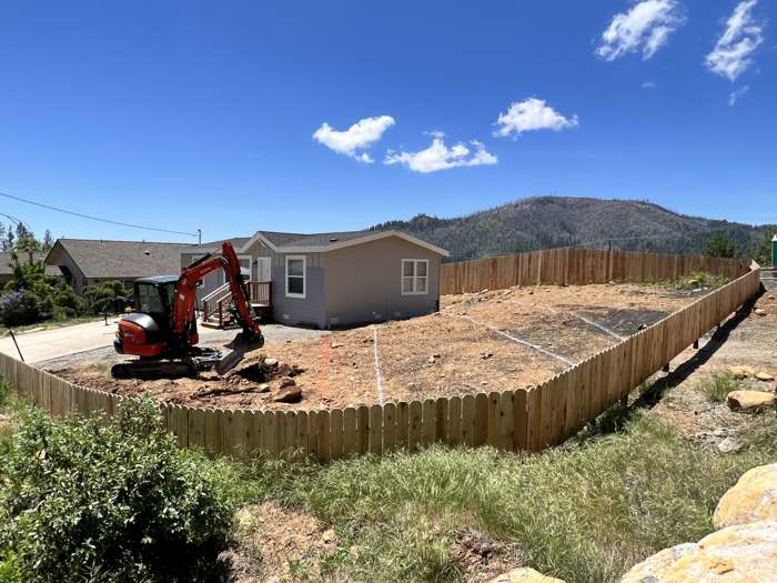R2's excavator grading a cleared lot beside a house, mountains behind