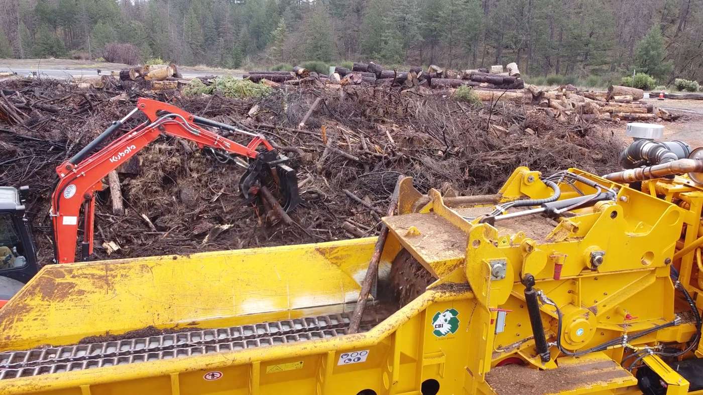 R2's excavator loading brush into the grinder, a yard of slash and logs behind
