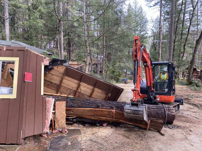 R2's excavator lifting a pine log off a crushed shed