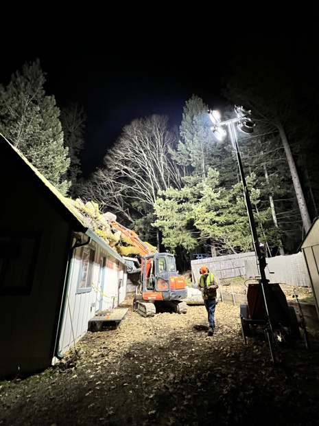 R2 working after dark under a light tower, the excavator beside a house with a tree on the roof